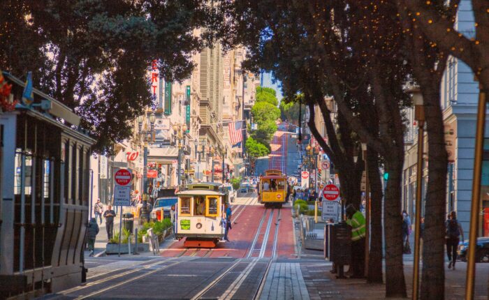 Street view of a city with two cable cars traveling down a hill, surrounded by buildings, trees with string lights, and pedestrians on the sidewalk—a charming scene often featured on the California Parks Highlights Tour from Los Angeles to San Francisco over 5 days.