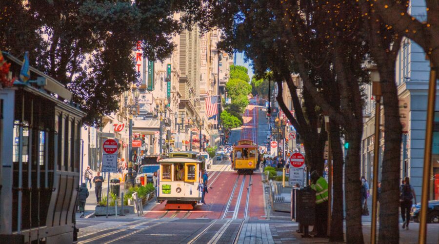 Street view of a city with two cable cars traveling down a hill, surrounded by buildings, trees with string lights, and pedestrians on the sidewalk—a charming scene often featured on the California Parks Highlights Tour from Los Angeles to San Francisco over 5 days.