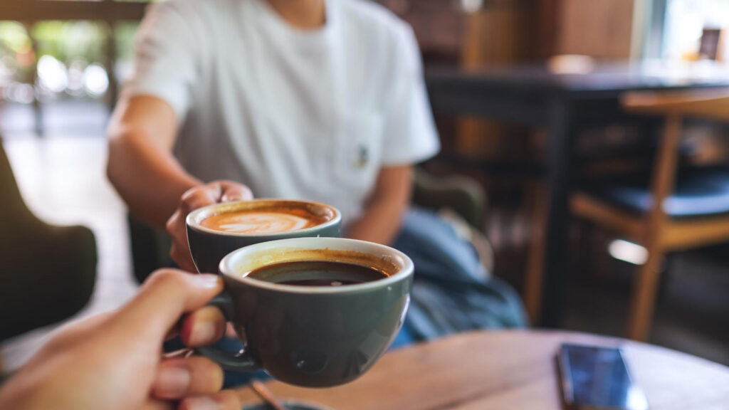 Two people wearing casual clothing clink ceramic mugs of coffee over a wooden table in a cozy San Francisco café setting.