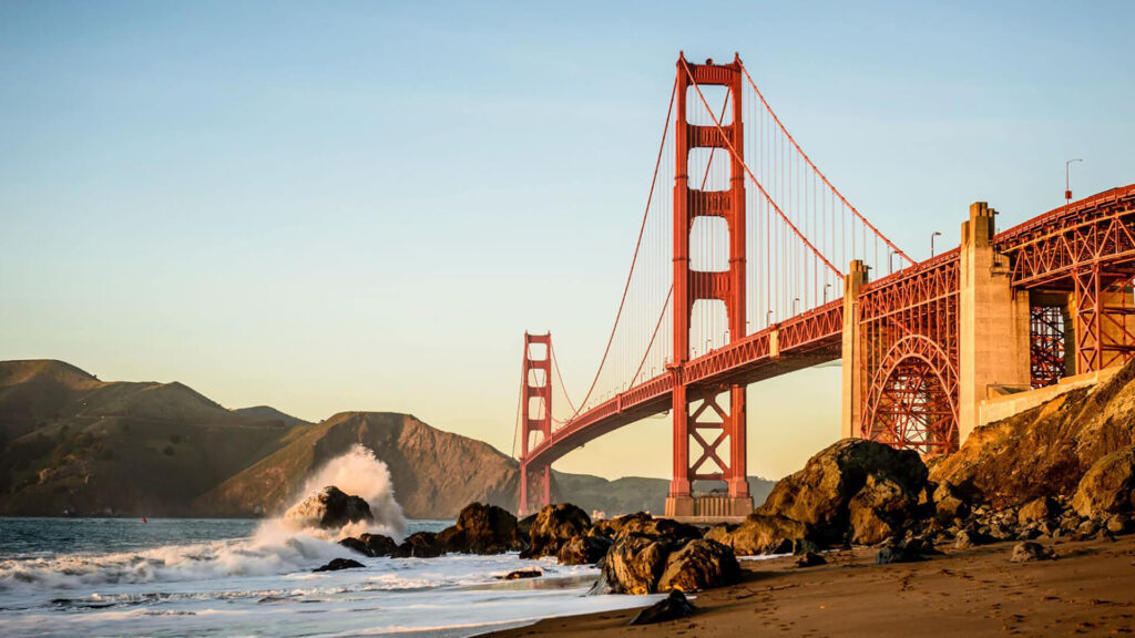 The Golden Gate Bridge stretches over a calm bay with waves crashing against rocks in the foreground and hills in the background, illuminated by warm sunlight.