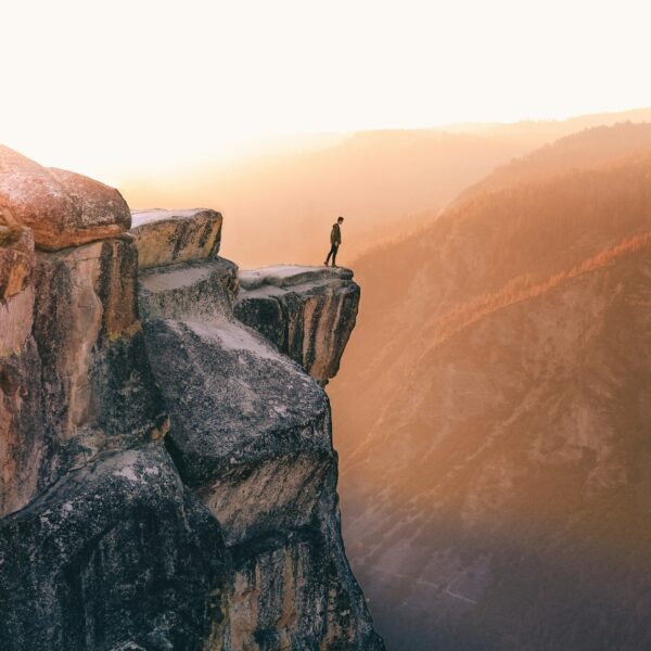 A person stands on the edge of a rocky cliff at sunset, overlooking the vast mountain landscape featured in the 5-day Highlights Tour of Yosemite.