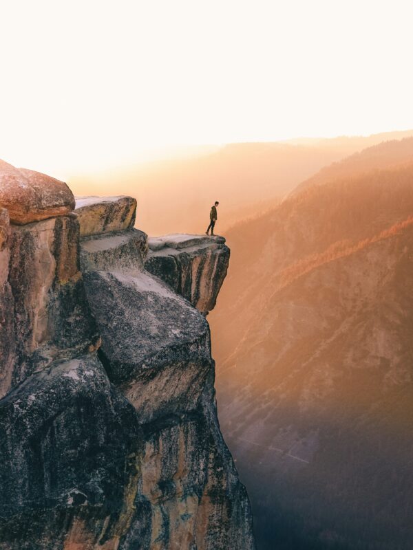 A person stands on the edge of a rocky cliff at sunset, overlooking the vast mountain landscape featured in the 5-day Highlights Tour of Yosemite.