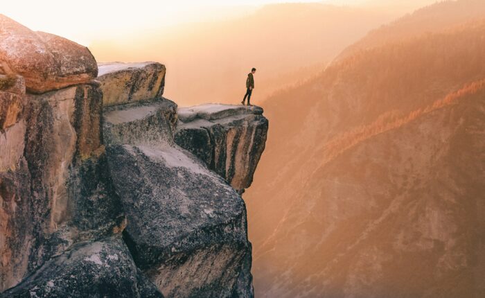 A person stands on the edge of a rocky cliff at sunset, overlooking the vast mountain landscape featured in the 5-day Highlights Tour of Yosemite.