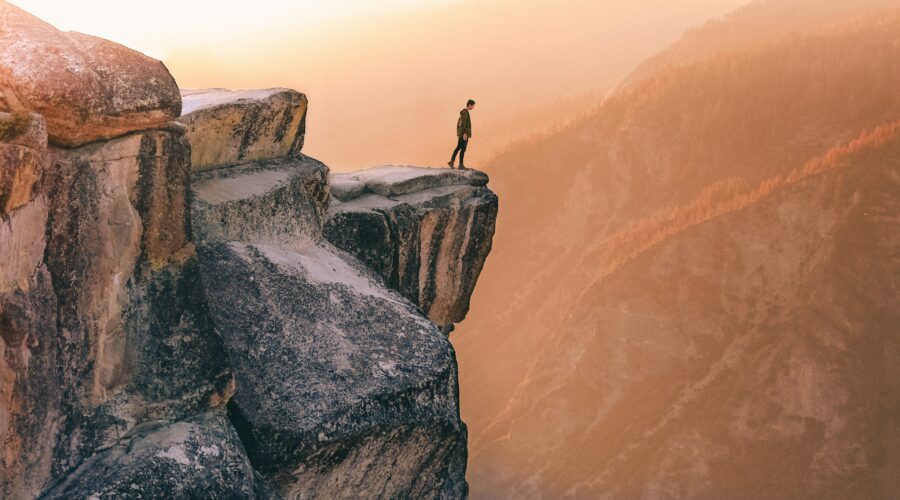 A person stands on the edge of a rocky cliff at sunset, overlooking the vast mountain landscape featured in the 5-day Highlights Tour of Yosemite.