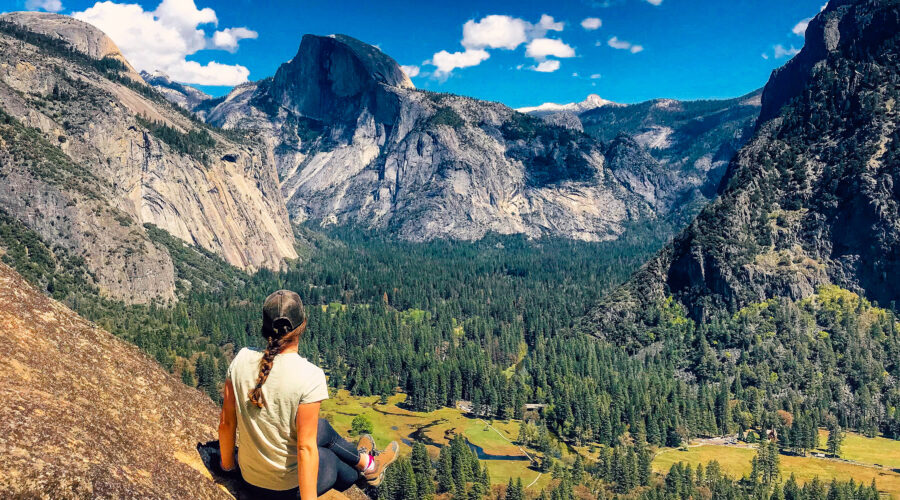 Person sitting on a rock ledge overlooking a vast mountainous landscape with forests and open fields under a blue sky, soaking in the breathtaking views on their Yosemite Highlights Tour. Perfect setting for a memorable 5-day adventure.