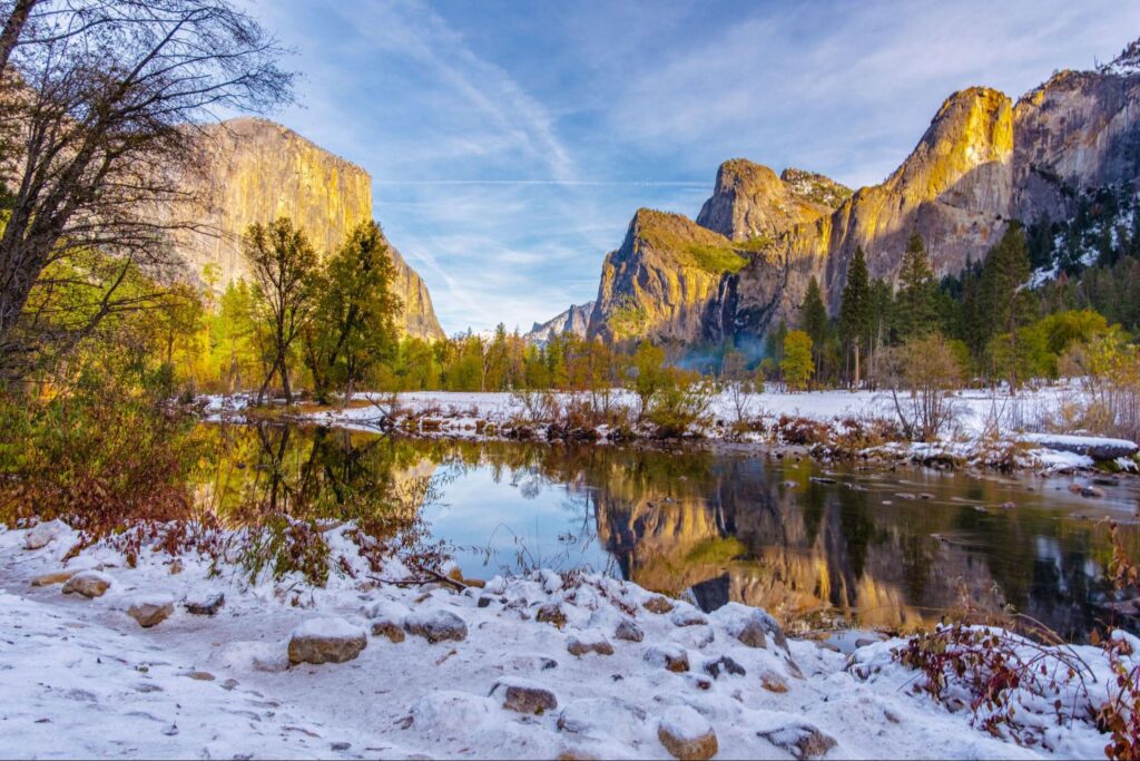 A river flows through a snowy valley with rocky cliffs and pine trees, reflecting the vibrant autumnal scenery and a bright sky.