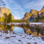 A river flows through a snowy valley with rocky cliffs and pine trees, reflecting the vibrant autumnal scenery and a bright sky.