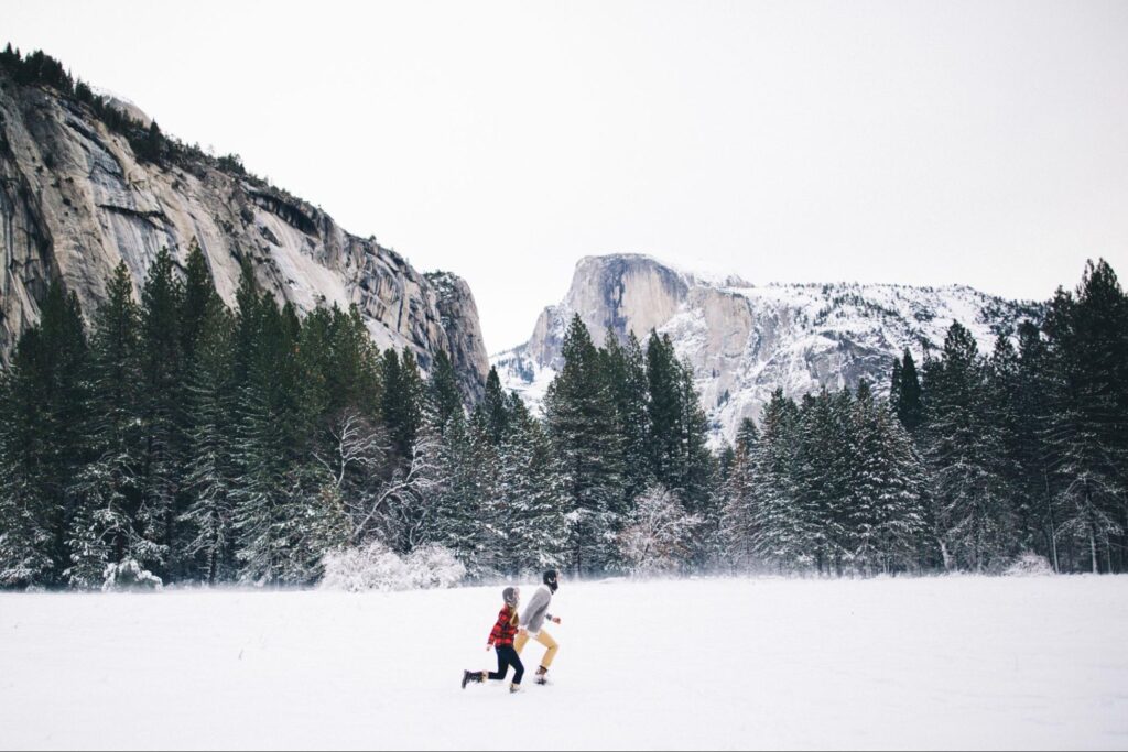 Two people play in a snowy meadow with mountains and evergreen trees in the background.