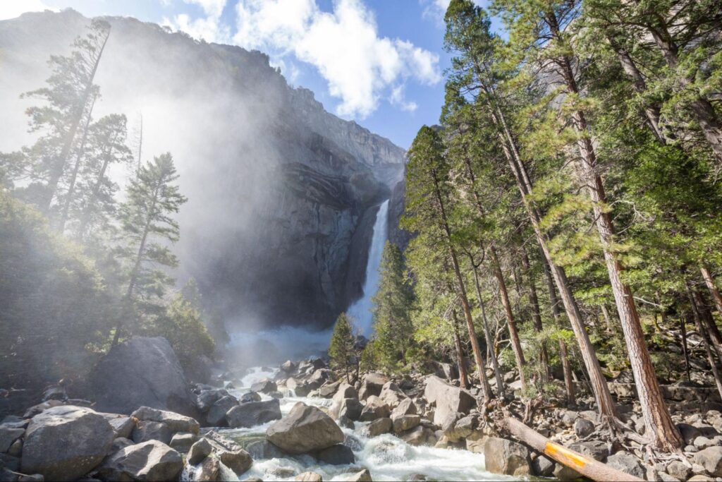 A column of water cascades down a rocky cliff surrounded by mist, trees, and boulders under a partly cloudy sky in a mountainous area.