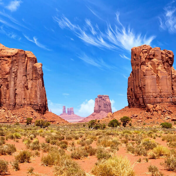 A desert landscape with large red rock formations under a bright blue sky with wispy clouds. Sparse vegetation covers the foreground.