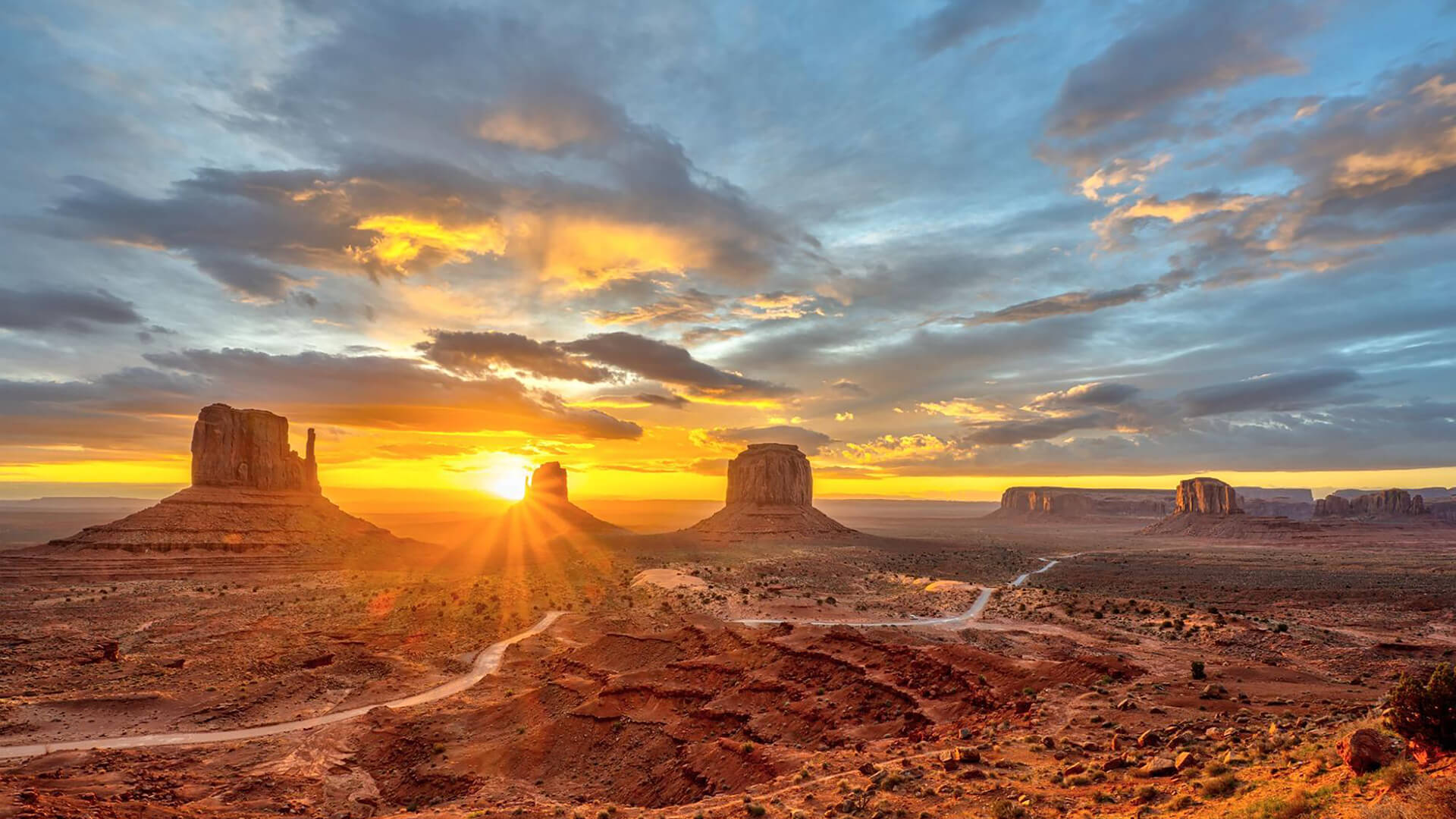 A desert landscape at sunrise featuring rock formations, rugged terrain, and a vast expanse of sky with clouds.