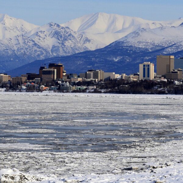 A city skyline with tall buildings stands in front of snowy mountains. The foreground features a frozen body of water with patches of ice.