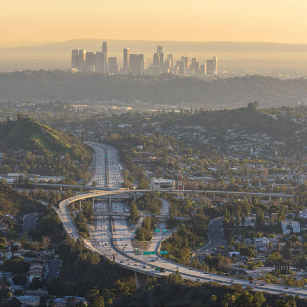 Aerial view of Los Angeles showcases its sprawling highways and lush green hills in the foreground, with the iconic city skyline visible under a characteristically hazy sky.