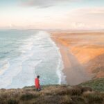 A person in a red jacket stands on a cliff overlooking a wide sandy beach with waves, reminiscent of the serene beauty found on a Muir Woods and Sausalito tour. The landscape is bathed in soft light under a partly cloudy sky.