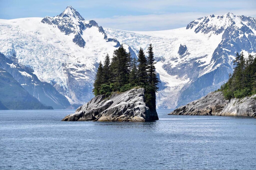 Rocky island with trees in a calm blue lake, surrounded by snow-capped mountains under a clear sky.