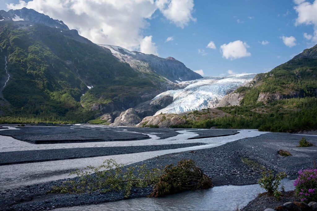 A glacier flows between green mountains under a blue sky with scattered clouds. A river winds through the rocky terrain in the foreground.