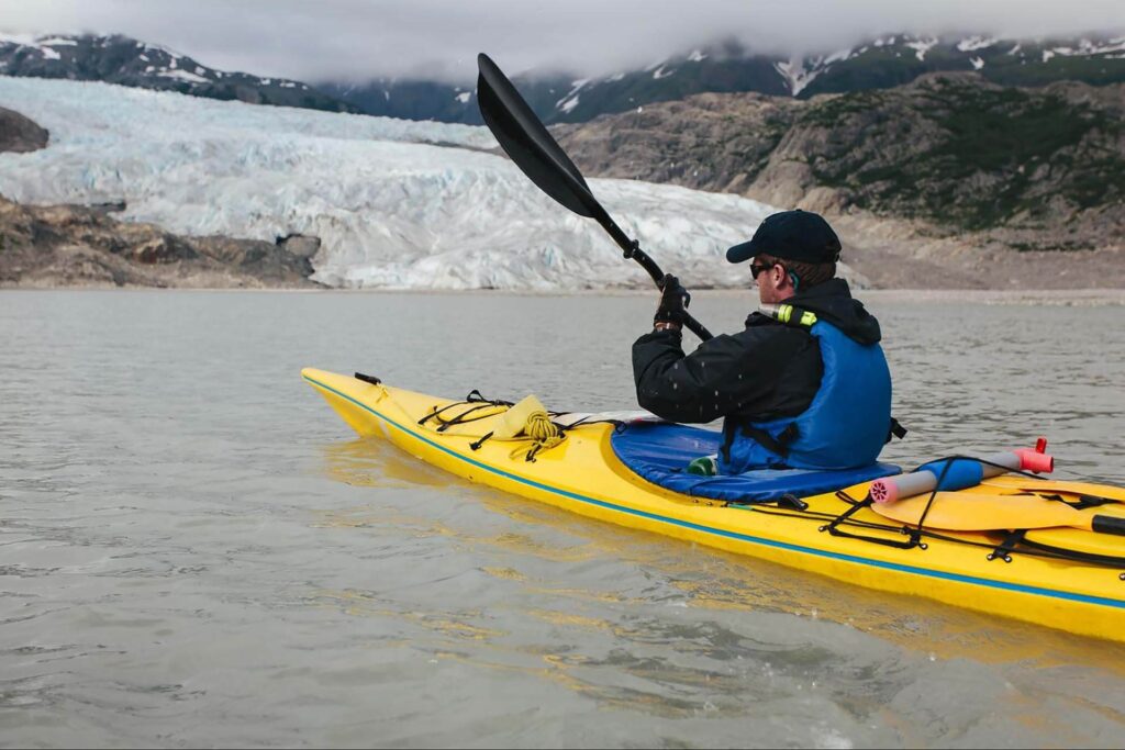 Person kayaking in a yellow kayak on a calm lake with a glacier and mountains in the background, under a cloudy sky.