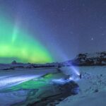 Under a starry sky, a person shines a flashlight towards the vibrant green northern lights near Anchorage, illuminating the snowy landscape and partially frozen river. It's a mesmerizing scene that captures the magic of an unforgettable tour in this winter wonderland.