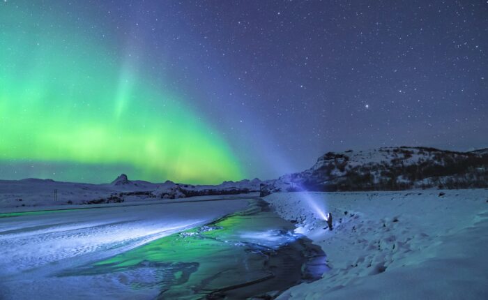 Under a starry sky, a person shines a flashlight towards the vibrant green northern lights near Anchorage, illuminating the snowy landscape and partially frozen river. It's a mesmerizing scene that captures the magic of an unforgettable tour in this winter wonderland.