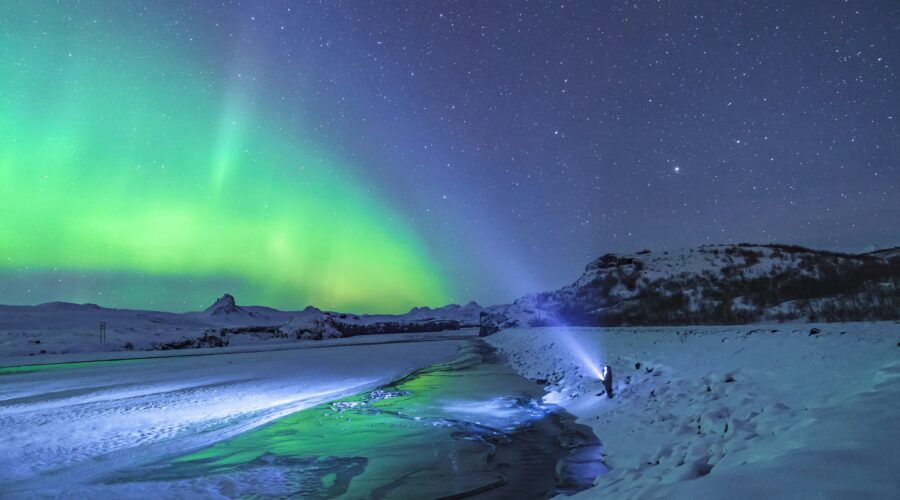 Under a starry sky, a person shines a flashlight towards the vibrant green northern lights near Anchorage, illuminating the snowy landscape and partially frozen river. It's a mesmerizing scene that captures the magic of an unforgettable tour in this winter wonderland.