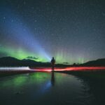 A person stands on a frozen landscape in Fairbanks, with a backdrop of the starry sky and Northern Lights, illuminated by colorful auroras on this mesmerizing tour.