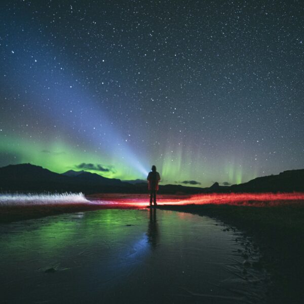 A person stands on a frozen landscape in Fairbanks, with a backdrop of the starry sky and Northern Lights, illuminated by colorful auroras on this mesmerizing tour.