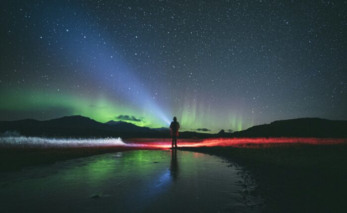 A person stands on a frozen landscape in Fairbanks, with a backdrop of the starry sky and Northern Lights, illuminated by colorful auroras on this mesmerizing tour.