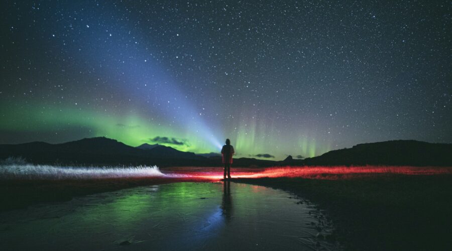 A person stands on a frozen landscape in Fairbanks, with a backdrop of the starry sky and Northern Lights, illuminated by colorful auroras on this mesmerizing tour.