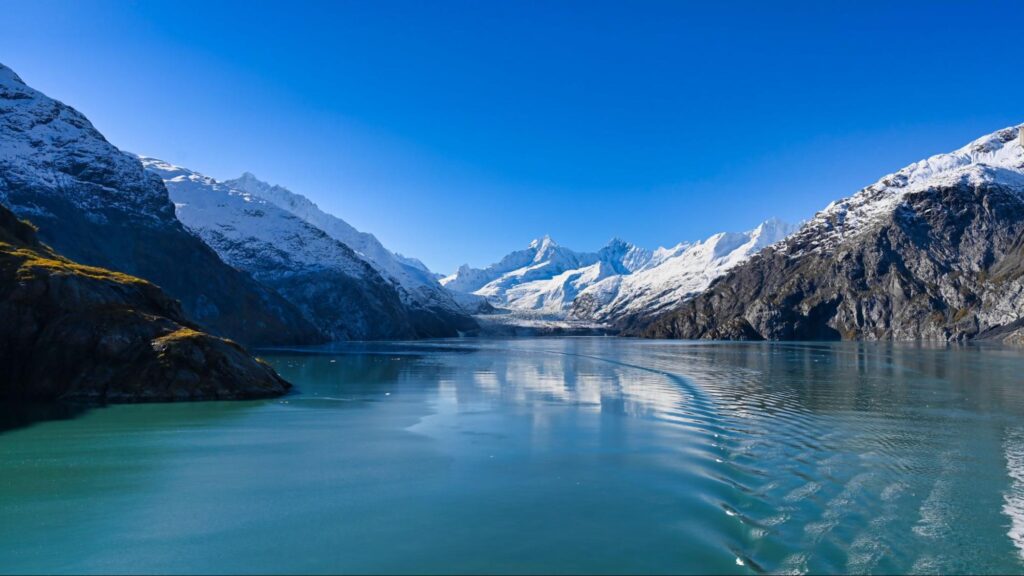 A serene glacier valley with clear blue water, surrounded by snow-capped mountains under a bright blue sky, offers the perfect backdrop for an unforgettable Alaska travel experience. Imagine exploring this breathtaking landscape where the essence of Denali meets the allure of Kenai Fjords.