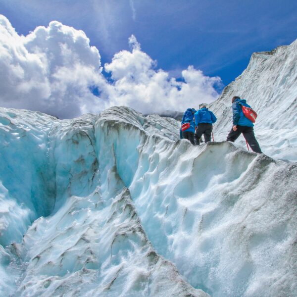 Three people in blue jackets hike up Matanuska Glacier's steep, icy slopes under a partly cloudy sky, embracing the thrill of their summer tour.