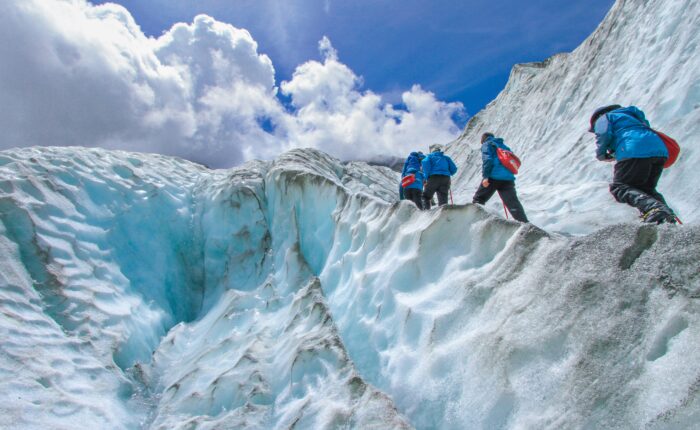 Three people in blue jackets hike up Matanuska Glacier's steep, icy slopes under a partly cloudy sky, embracing the thrill of their summer tour.