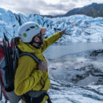 A person in a yellow jacket and helmet points towards Matanuska Glacier, equipped with a backpack and climbing gear. Other adventurers are visible in the distance on this exhilarating winter tour across the icy terrain.