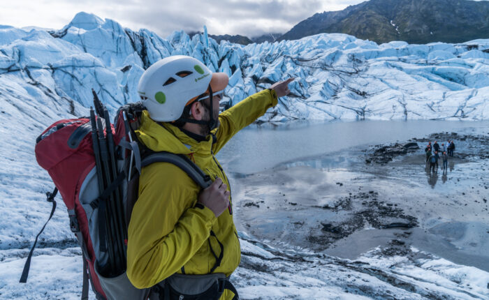 A person in a yellow jacket and helmet points towards Matanuska Glacier, equipped with a backpack and climbing gear. Other adventurers are visible in the distance on this exhilarating winter tour across the icy terrain.