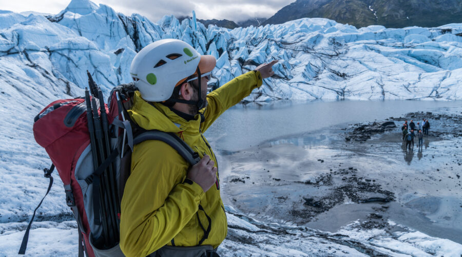 A person in a yellow jacket and helmet points towards Matanuska Glacier, equipped with a backpack and climbing gear. Other adventurers are visible in the distance on this exhilarating winter tour across the icy terrain.