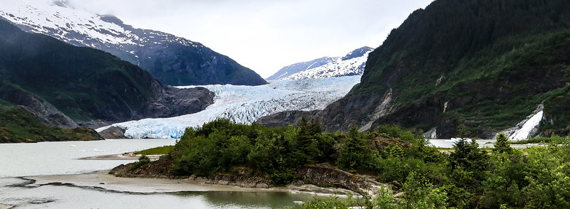 A glacier of blue and white ice stretches majestically between two forested mountains in Kenai Fjords, with a serene body of water in the foreground. The overcast sky adds a mysterious allure, inviting Alaska travel enthusiasts to explore this breathtaking landscape.