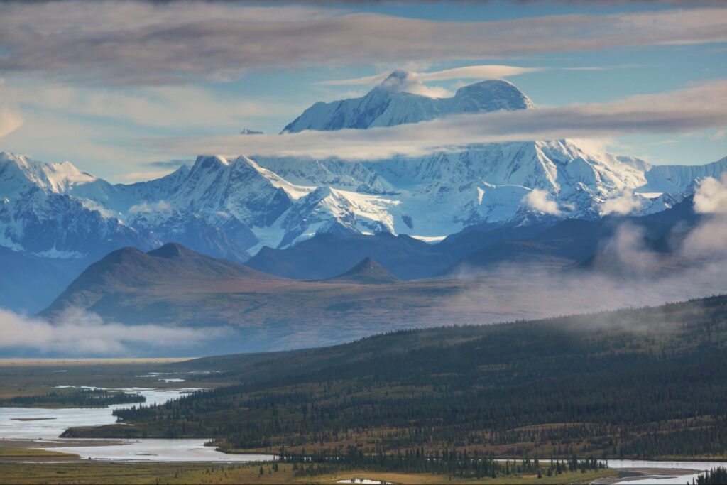 Denali's snow-capped mountain range rises majestically under a partly cloudy sky, with a river meandering through the lush forest in the foreground, creating an enchanting scene perfect for Alaska travel enthusiasts.