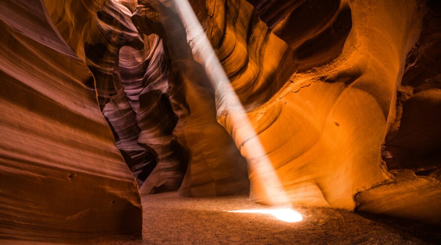 A beam of sunlight illuminates the sandy floor of a narrow, winding slot canyon with smooth, curved orange and purple rock walls