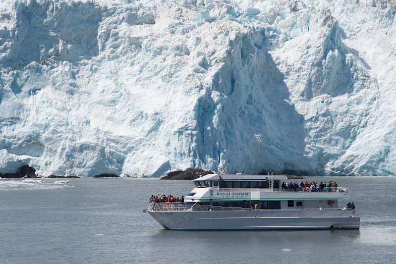 A tour boat with passengers glides through the serene waters of Resurrection Bay, offering breathtaking views as it approaches a massive glacier, showcasing the stunning beauty of Kenai Fjords.