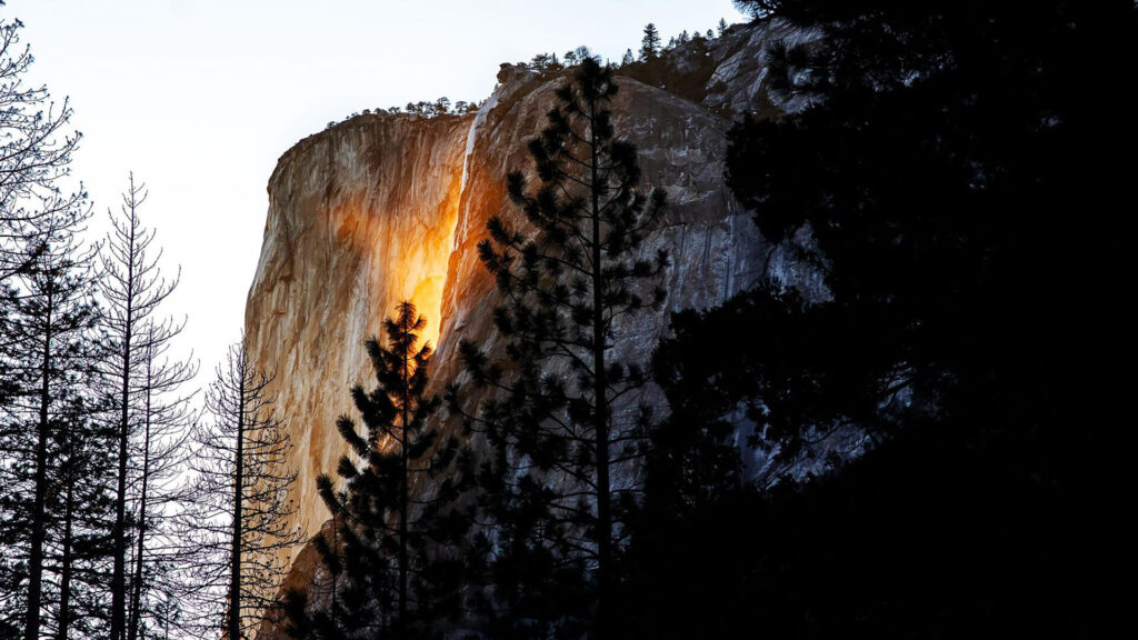 A rocky cliff is illuminated by sunlight, creating a glowing effect reminiscent of the 2025 Yosemite Firefall, while a cascade streams gracefully on the left side, surrounded by silhouetted trees.