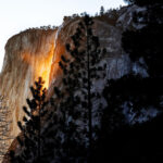 A rocky cliff is illuminated by sunlight, creating a glowing effect reminiscent of the 2025 Yosemite Firefall, while a cascade streams gracefully on the left side, surrounded by silhouetted trees.