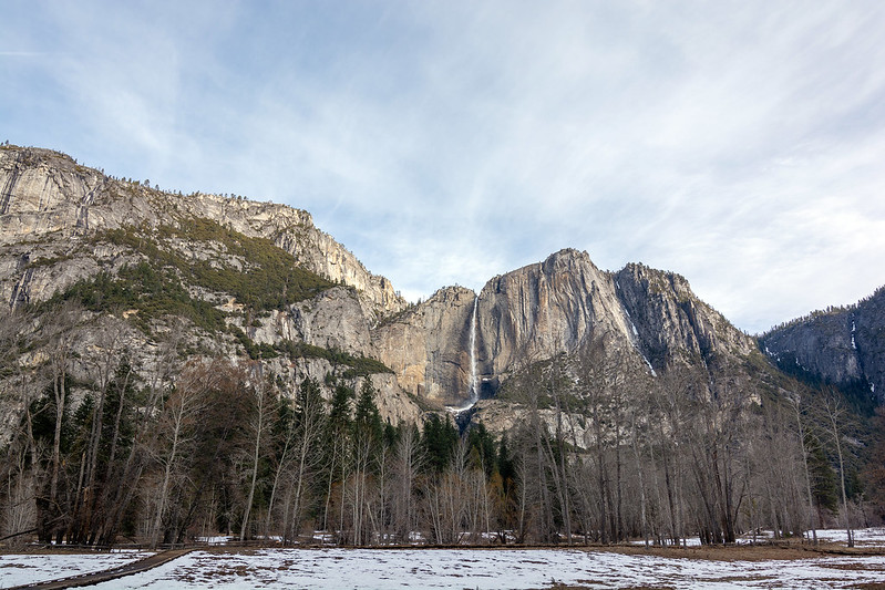 A landscape view of rocky mountains, with sparse snow and trees at the base, stretches beneath a partly cloudy sky. This scene evokes memories of the famed Yosemite Firefall and promises incredible adventures for those exploring these majestic terrains in 2025.