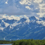 Snow-capped mountains under a cloudy sky, with green foliage and a river in the foreground, capture the essence of an Alaska road trip from Anchorage to Seward.