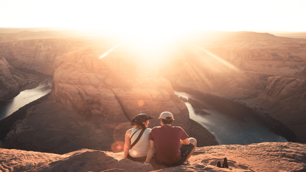 A couple sits on a cliff edge overlooking Horseshoe Bend at sunset, with the Colorado River visible below and sun rays casting light over the scene.