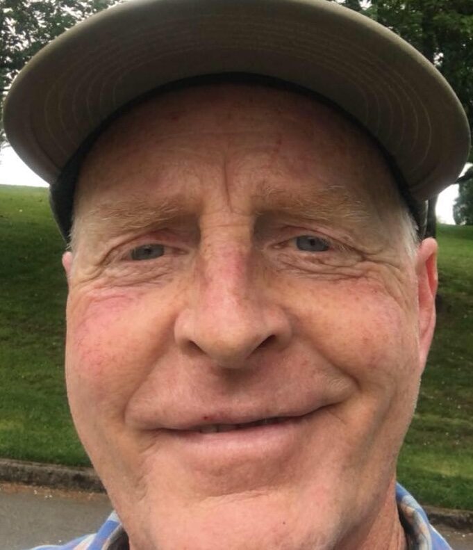 A person wearing a cap smiles at the camera with grass and trees blurred in the background.