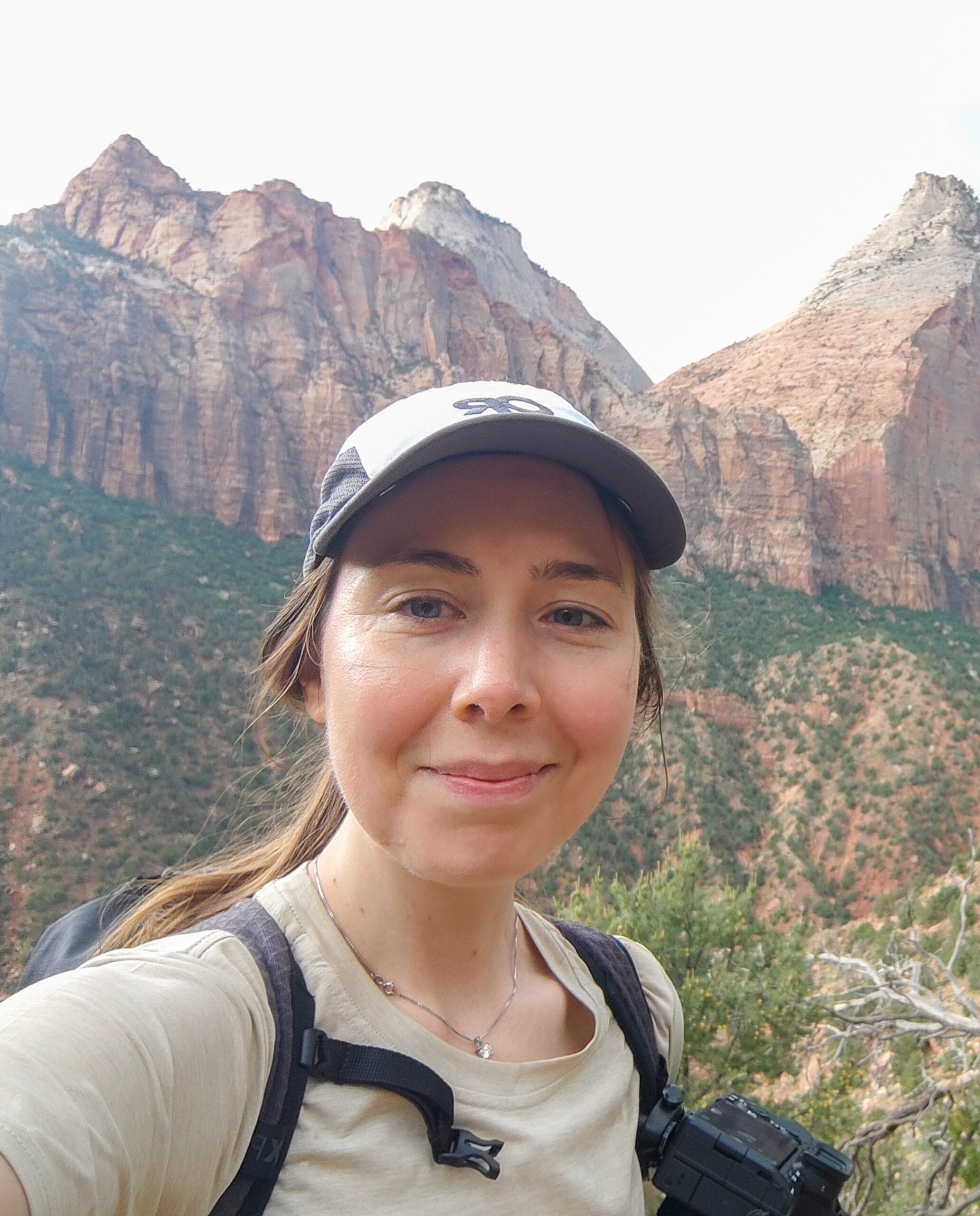 Hannah Freeman, sporting a cap and backpack, beams for a selfie against the backdrop of rugged mountainous terrain and sparse vegetation.