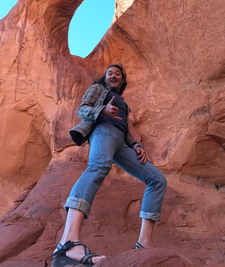 Person posing with a thumbs-up in front of rocky arch, wearing a plaid shirt, jeans, and sandals.