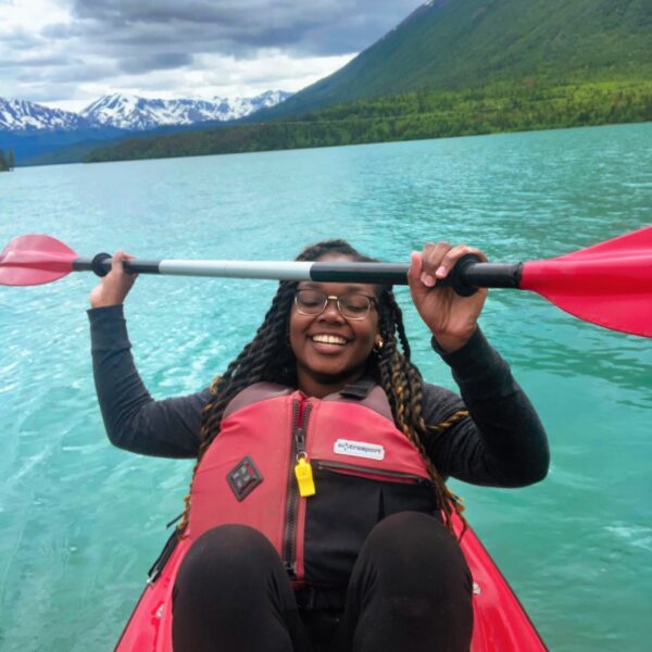 Kika Grimes-Boles, in a red life jacket, holds a paddle while sitting in a kayak on a turquoise lake. The scene is set against majestic mountains and lush trees under a softly cloudy sky.