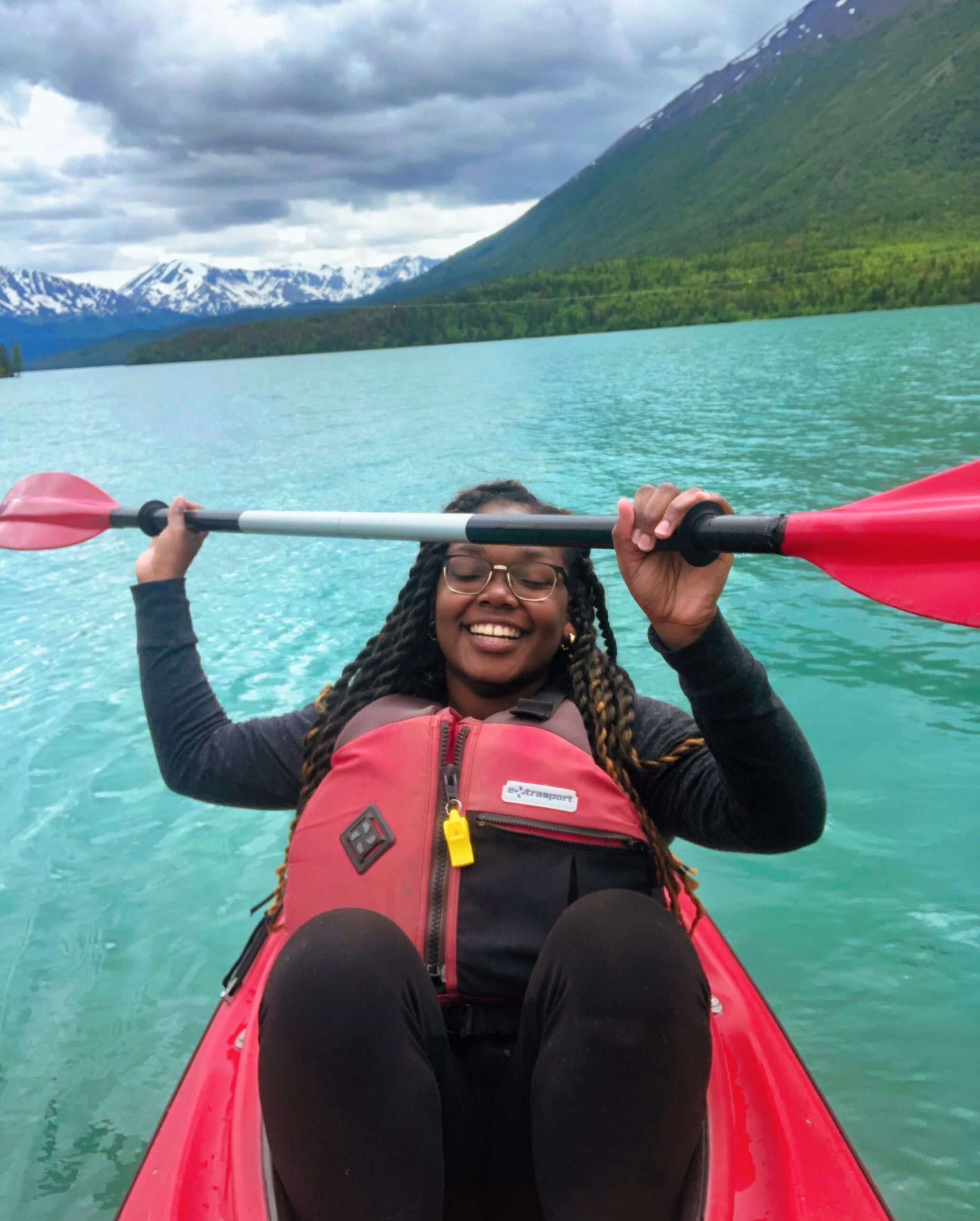 Kika Grimes-Boles, in a red life jacket, holds a paddle while sitting in a kayak on a turquoise lake. The scene is set against majestic mountains and lush trees under a softly cloudy sky.