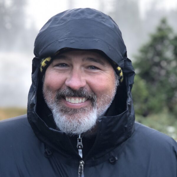 A smiling person, possibly inspired by a Jay Burleson film, wears a black hooded jacket, embracing the mysterious allure of the misty day outdoors.