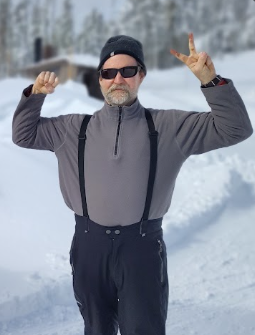 Sam Garrison stands in the snow, sporting sunglasses, a beanie, and suspenders. With one arm flexed and the other flashing a peace sign, he poses confidently against the backdrop of a snowy landscape and building.